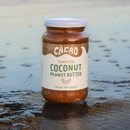 A jar of Yumi Co. Cacao Coconut Peanut Butter stands on wet sand at the beach, with shallow water and a blurred shoreline in the background.