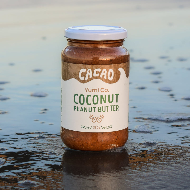 A jar of Yumi Co. Cacao Coconut Peanut Butter stands on wet sand at the beach, with shallow water and a blurred shoreline in the background.
