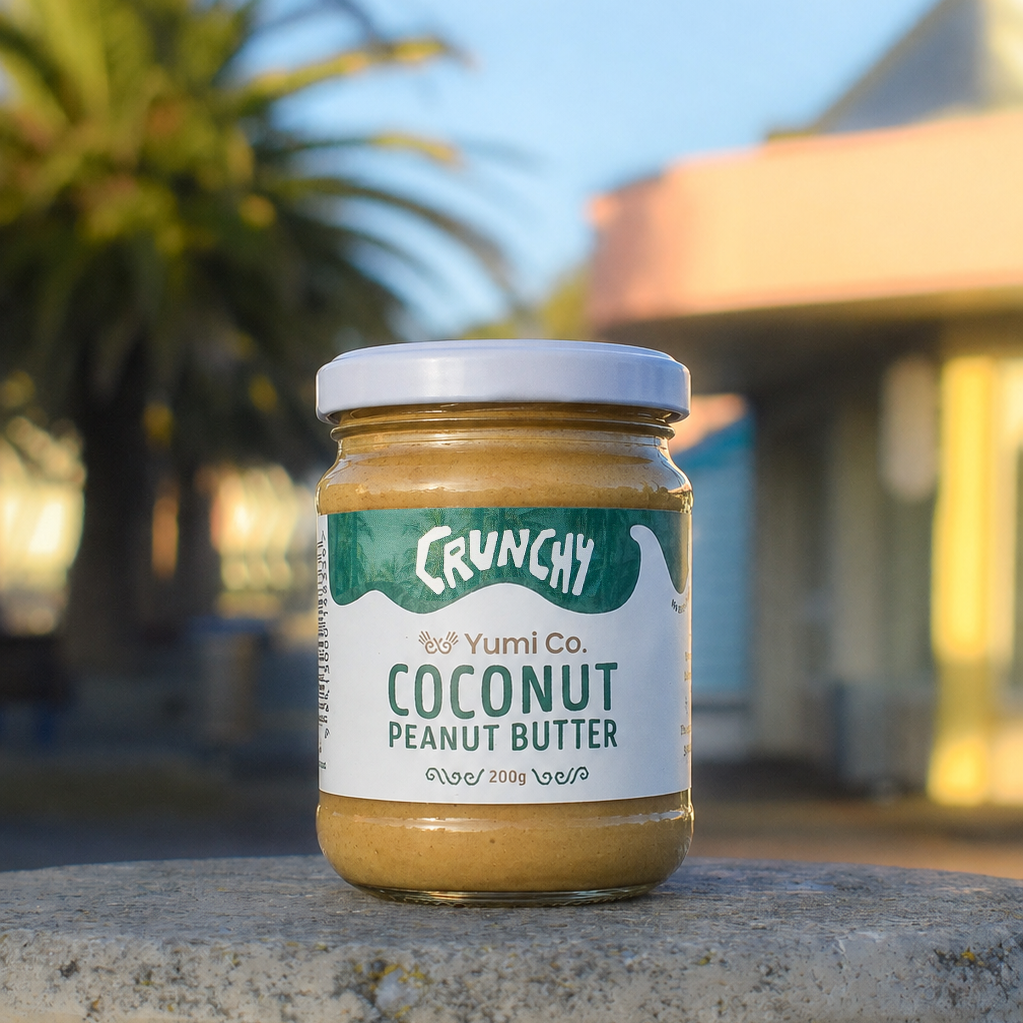 A jar of Crunchy Yumi Co. Coconut Peanut Butter sits on a concrete ledge outdoors with a blurred background of palm trees and a building.