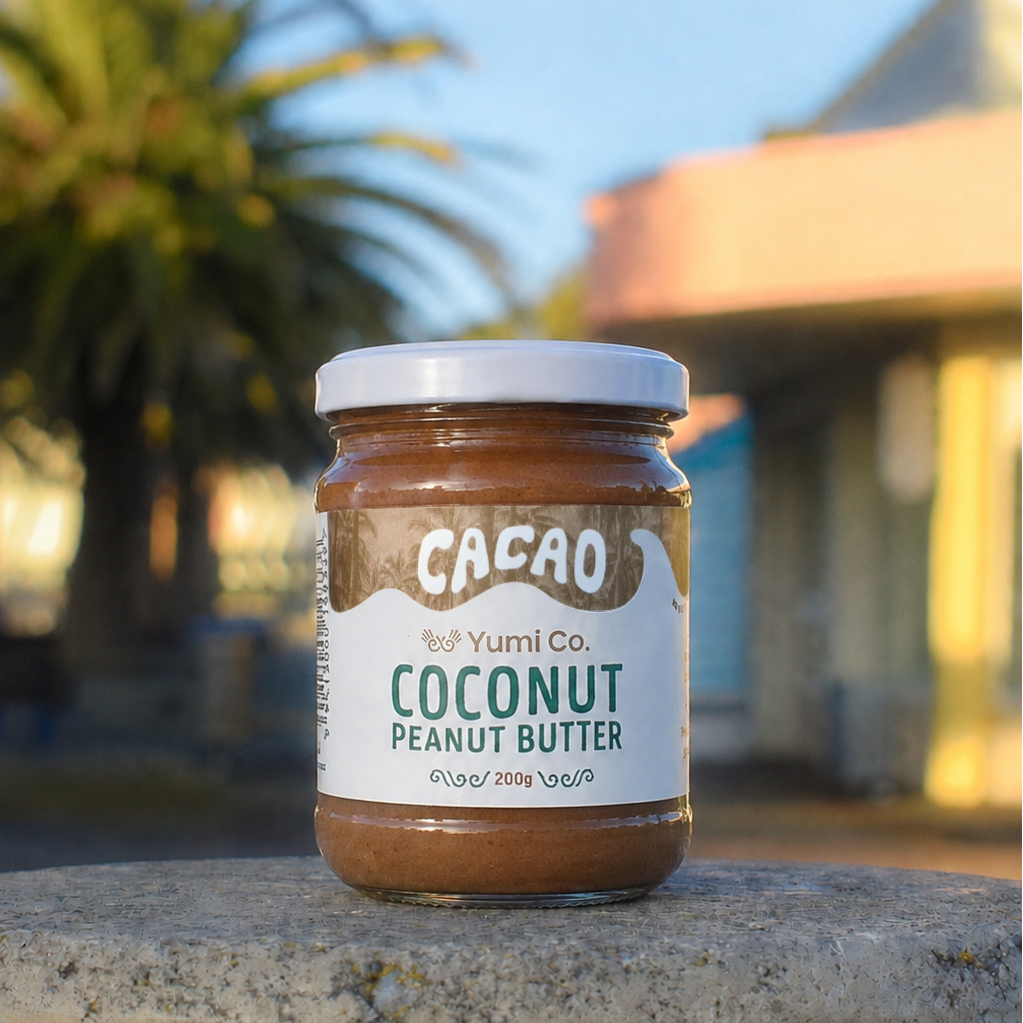 A jar of Yumi Co. Cacao Coconut Peanut Butter sits on a concrete ledge outdoors with a blurred background of palm trees and a building under a blue sky.