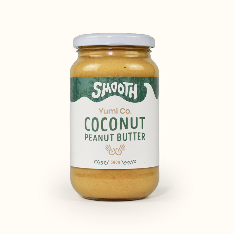 A glass jar of Yumi Co. Smooth Coconut Peanut Butter with a white lid and a white label featuring green and brown text, set against a solid off-white background.